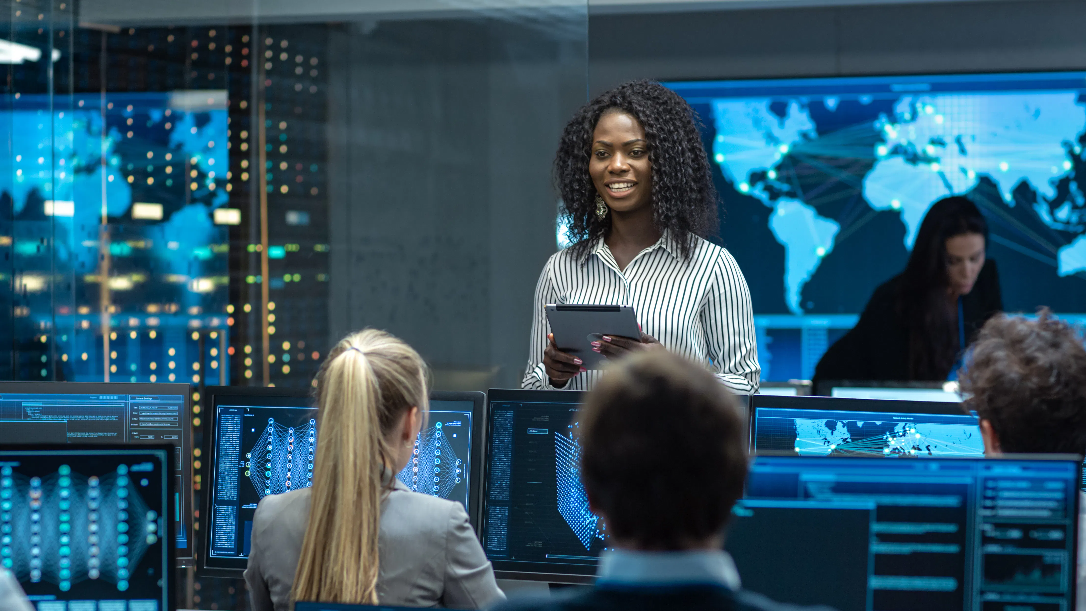 A machine learning engineer holding a tablet is briefing team members who are working on computers.